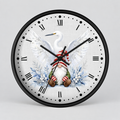 A round wall clock with a black frame and a white clock face featuring a festive illustration of a white heron and a Christmas gnome surrounded by blue winter foliage.