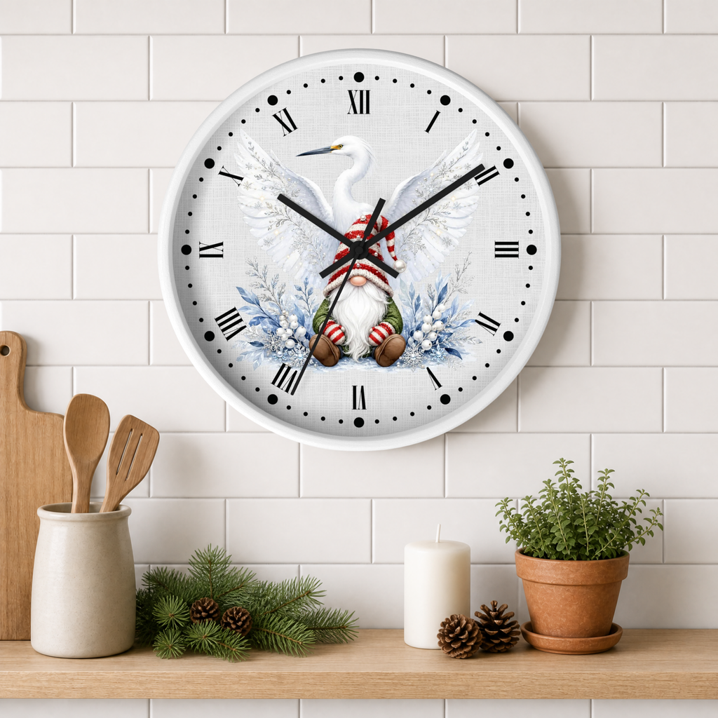 A round wall clock with a white face and Roman numerals, featuring a winter illustration of a white heron with wings spread, blue floral accents, and a small gnome in red-and-white striped pants. The clock is mounted on a white subway-tile wall above a wooden shelf styled with kitchen utensils, pine branches, a candle, and a potted plant.