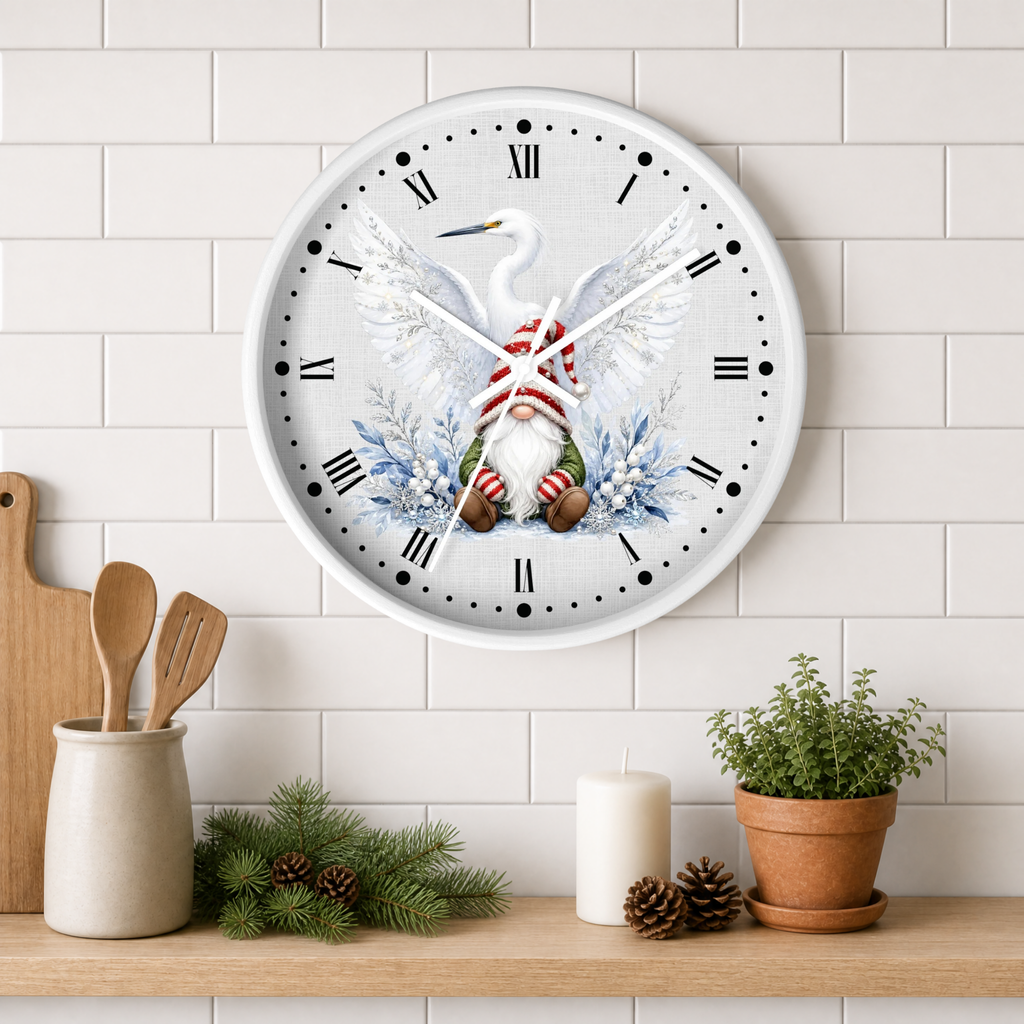 A round wall clock with a white face and Roman numerals, featuring a festive illustration of a white heron with spread wings, a red-and-white striped scarf, and a small gnome with a red beard and striped socks, surrounded by blue mist and evergreen branches. The clock is mounted on a white subway tile wall above a wooden shelf styled with kitchen utensils, pinecones, a candle, and a potted plant.