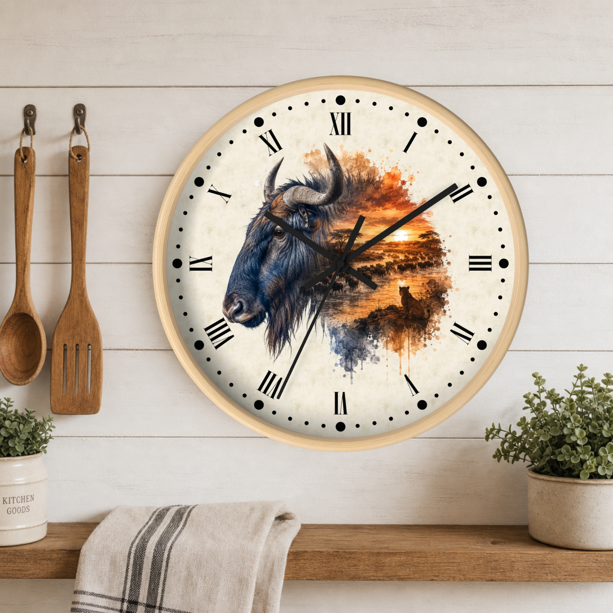 Round wall clock with a light wood frame and Roman numerals, featuring a watercolor-style Wildebeest illustration over an orange sunset scene. The clock is mounted on a white wood-paneled wall above a shelf with kitchen utensils, plants, and a towel.