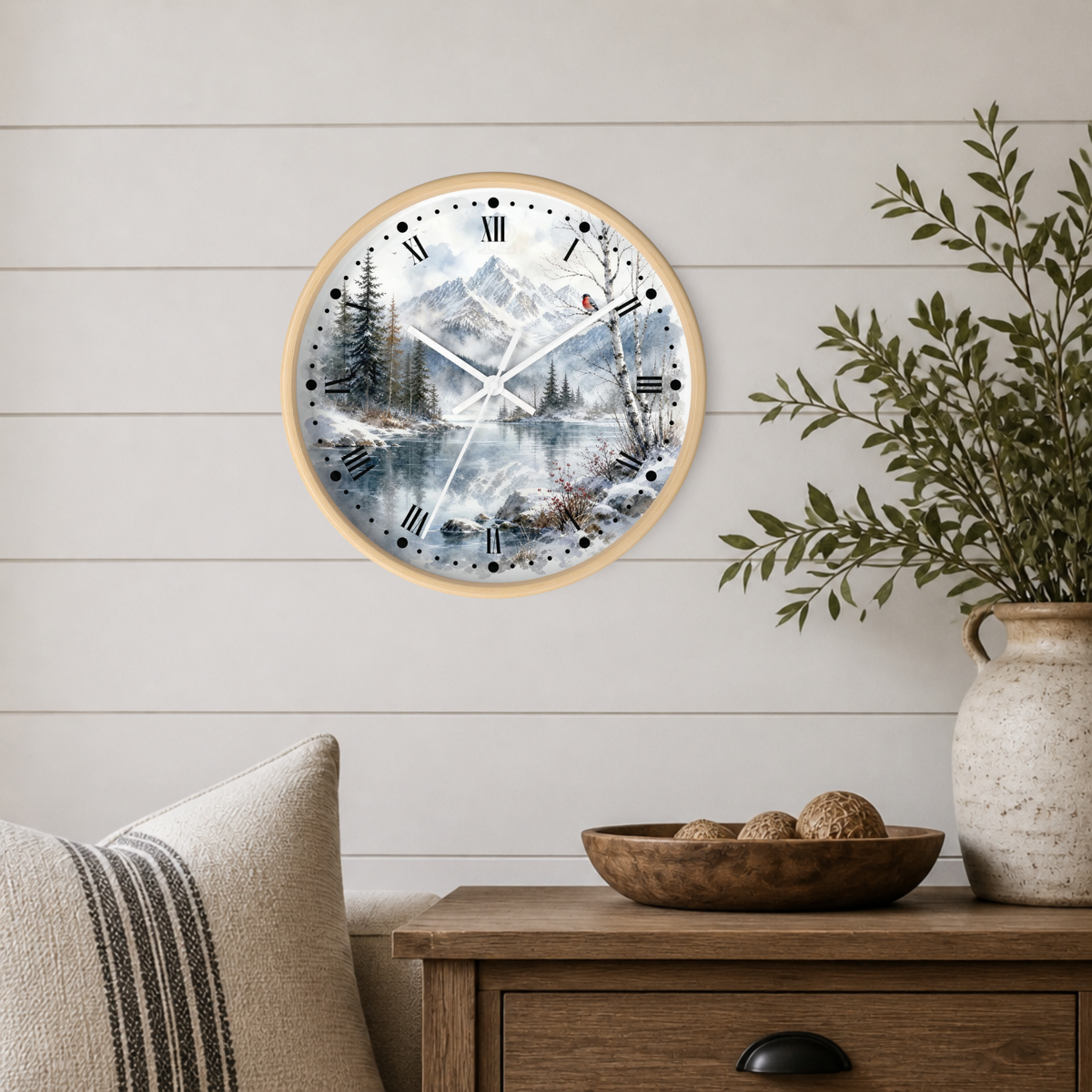 A round wall clock with a light wood frame and a scenic winter mountain landscape print, mounted on a white shiplap wall above a wooden side table with a plant and decorative bowl.