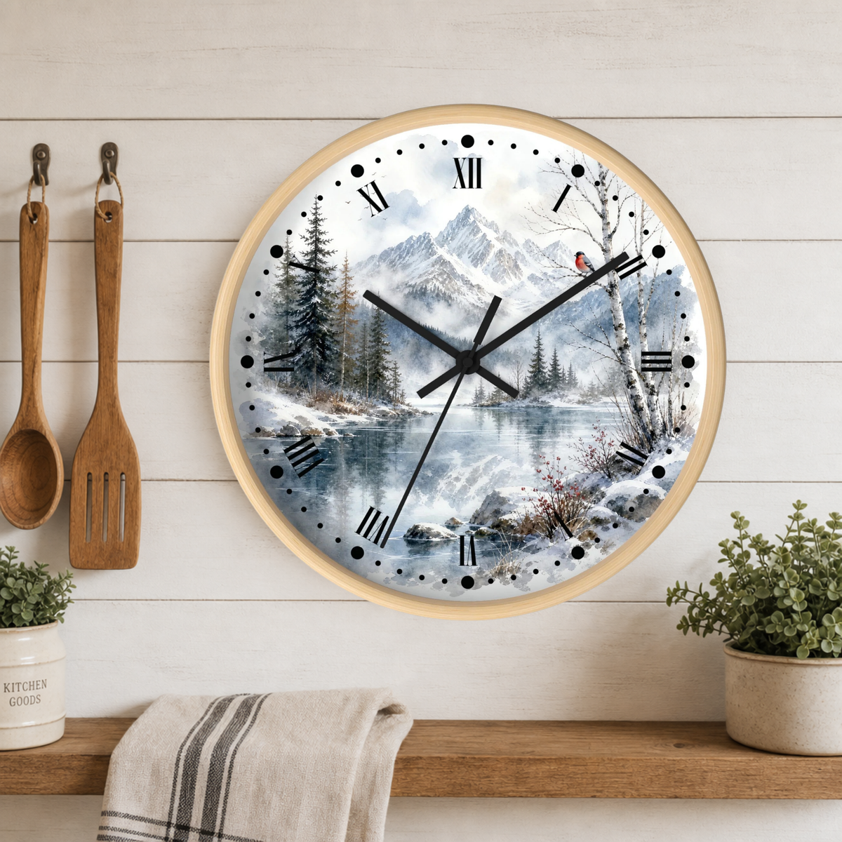 Round wall clock with a light wood frame and a scenic winter landscape print featuring snow-covered mountains, pine trees, and a reflective lake. The clock is mounted on a white wood-paneled wall above a wooden shelf with small potted plants and a towel, with wooden kitchen utensils hanging on the left.