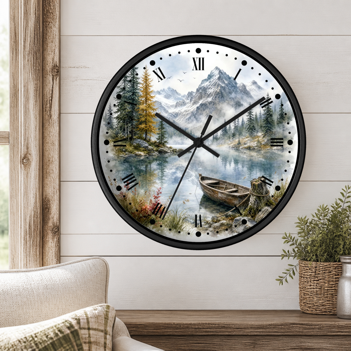 A round wall clock with a black frame and a scenic landscape print of a mountain lake, pine trees, and a rowboat. The clock is mounted on a white wood-paneled wall above a wooden shelf with a potted plant and part of a sofa visible.
