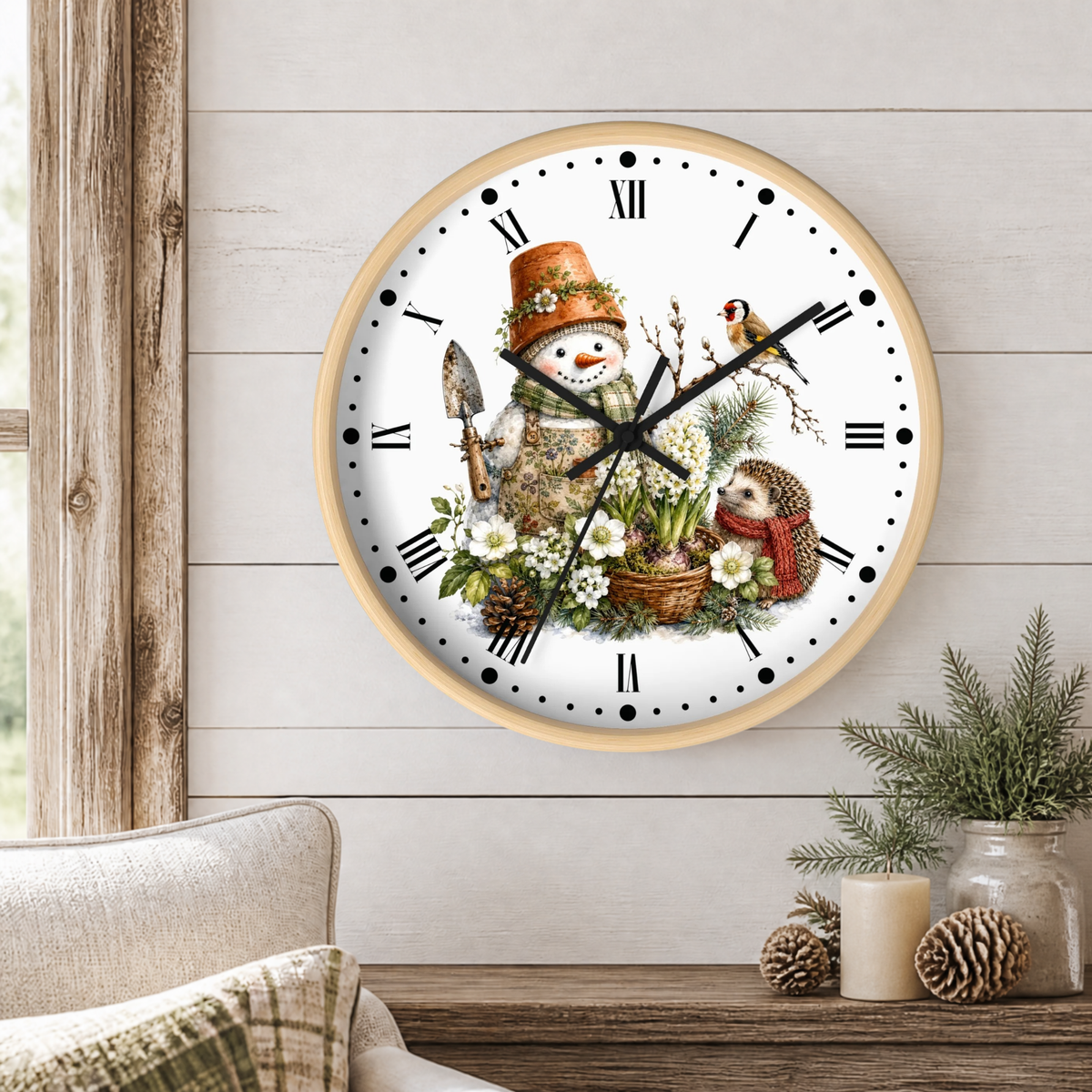 A round wall clock with a light wood frame and a whimsical winter garden illustration on the face, mounted on a white shiplap wall above a rustic wooden table with candles and pinecones.