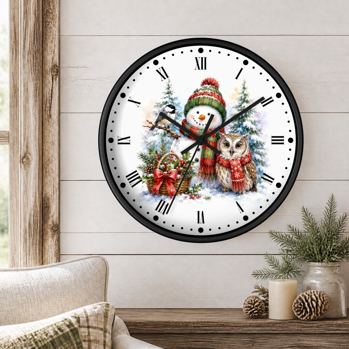 A round wall clock with a black frame and a festive winter illustration featuring a snowman in a red knit hat and scarf, an owl in a red scarf, Christmas trees, and ornaments. The clock is mounted on a light wood-paneled wall above a wooden table with a candle, pinecones, and a small plant, with a beige armchair partially visible on the left.