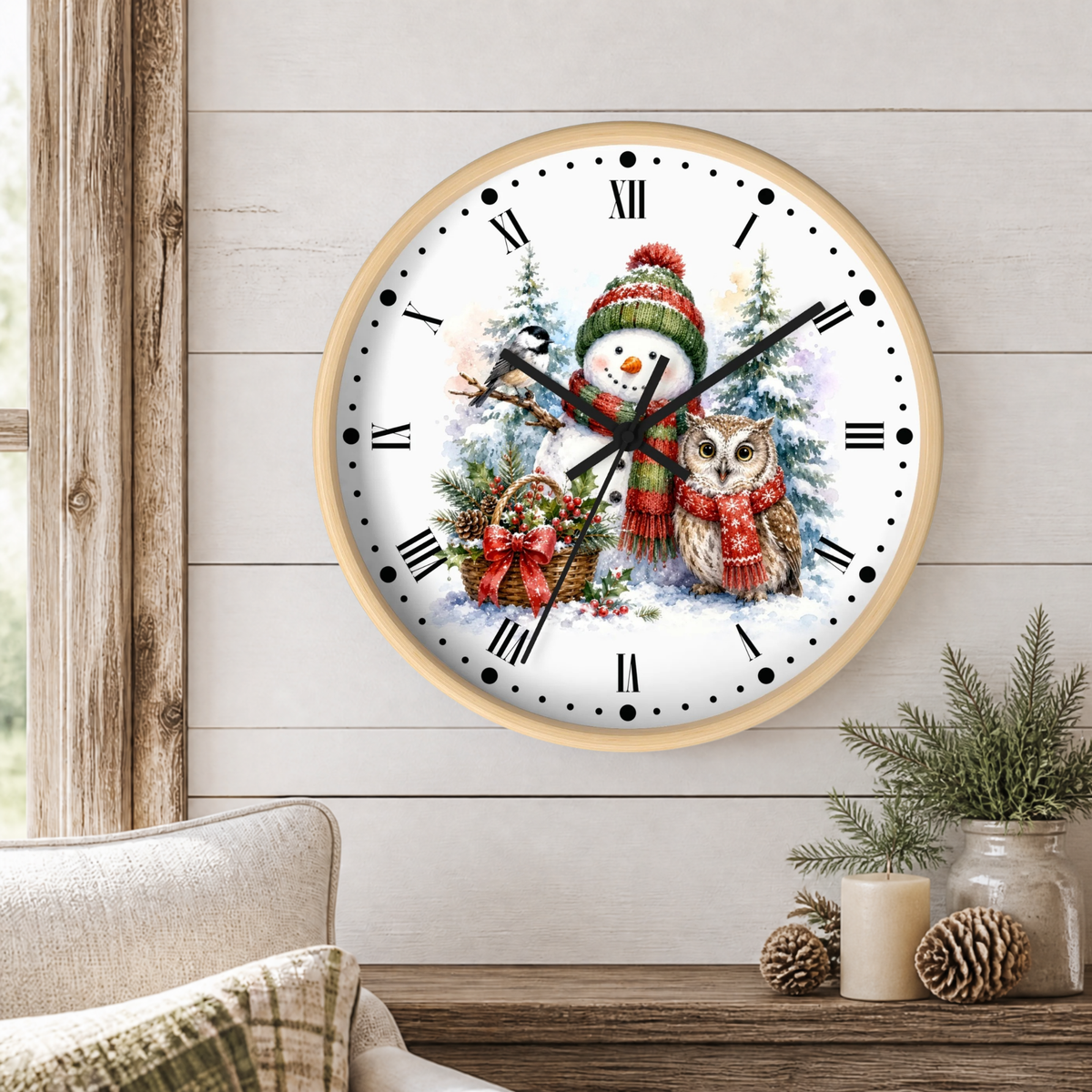 A round wall clock with a light wood frame and a festive winter illustration featuring a snowman, owl, evergreen trees, and a basket of ornaments. The clock is mounted on a white wood-paneled wall above a rustic wooden table with a candle, pinecones, and a small plant, with a beige armchair partially visible on the left.
