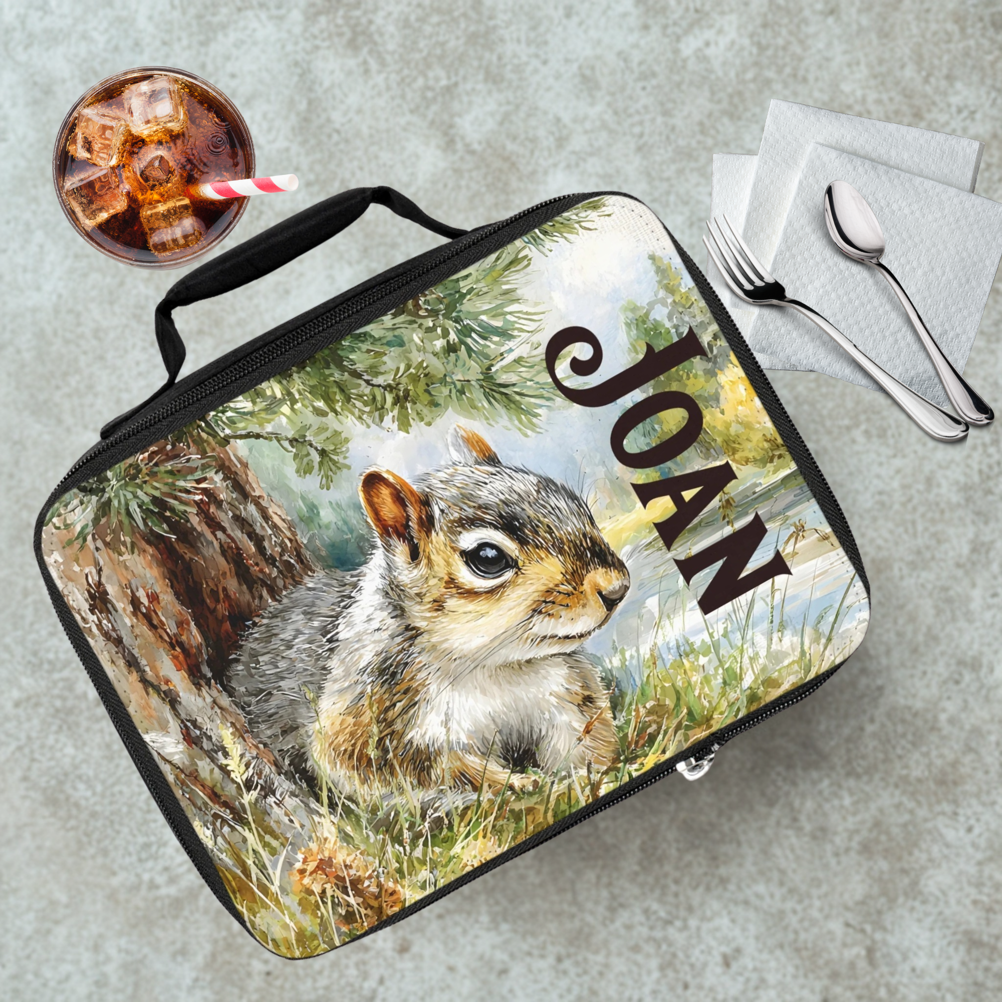 Lunch bag with a squirrel design and 'Joan' text on a cafeteria table with a drink and utensils.