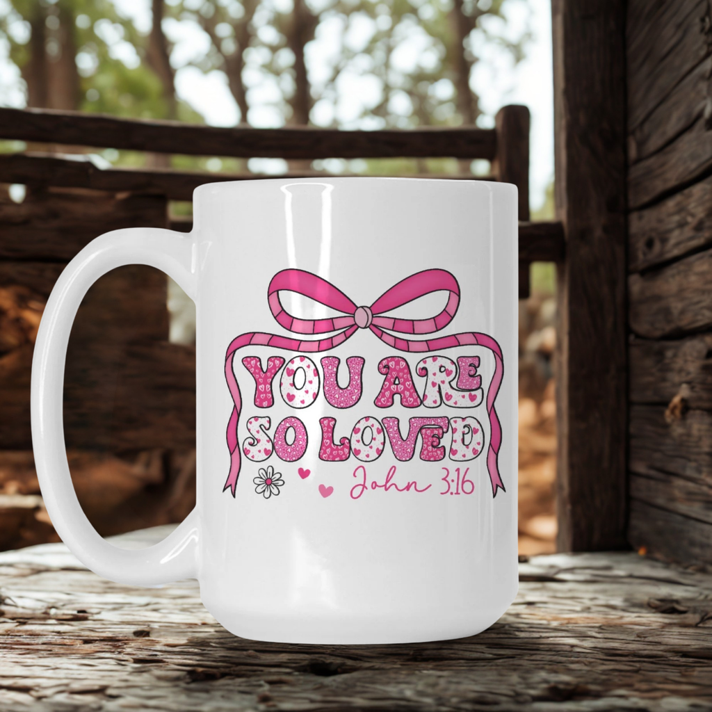 White mug with pink bow and text 'You are so loved John 3:16' on a wooden surface.