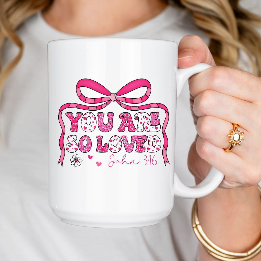 White mug with pink bow and text 'You are so loved John 3:16' held by a person.