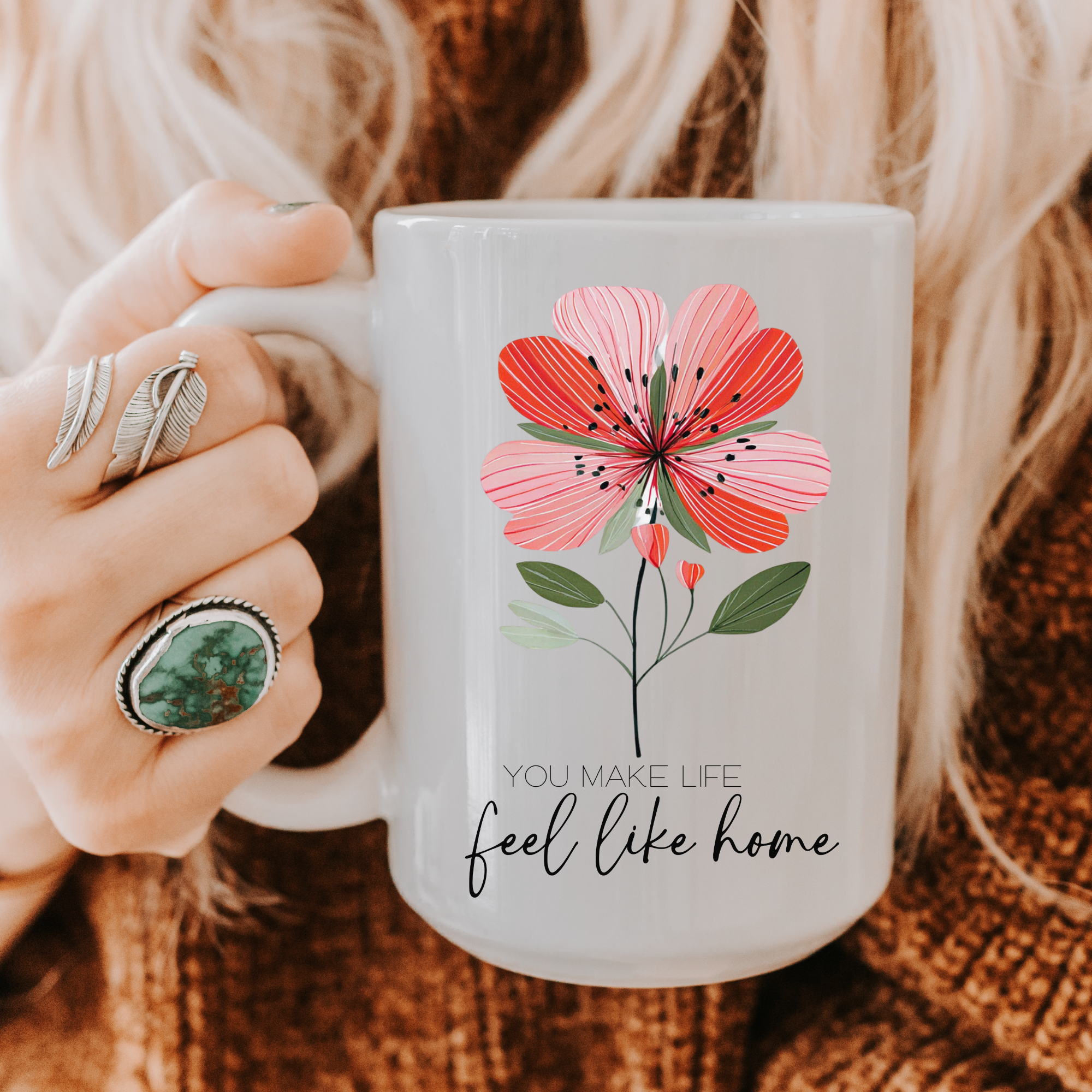 Person holding a 15oz. mug with a floral design and text 'You make life feel like home' on a textured surface.