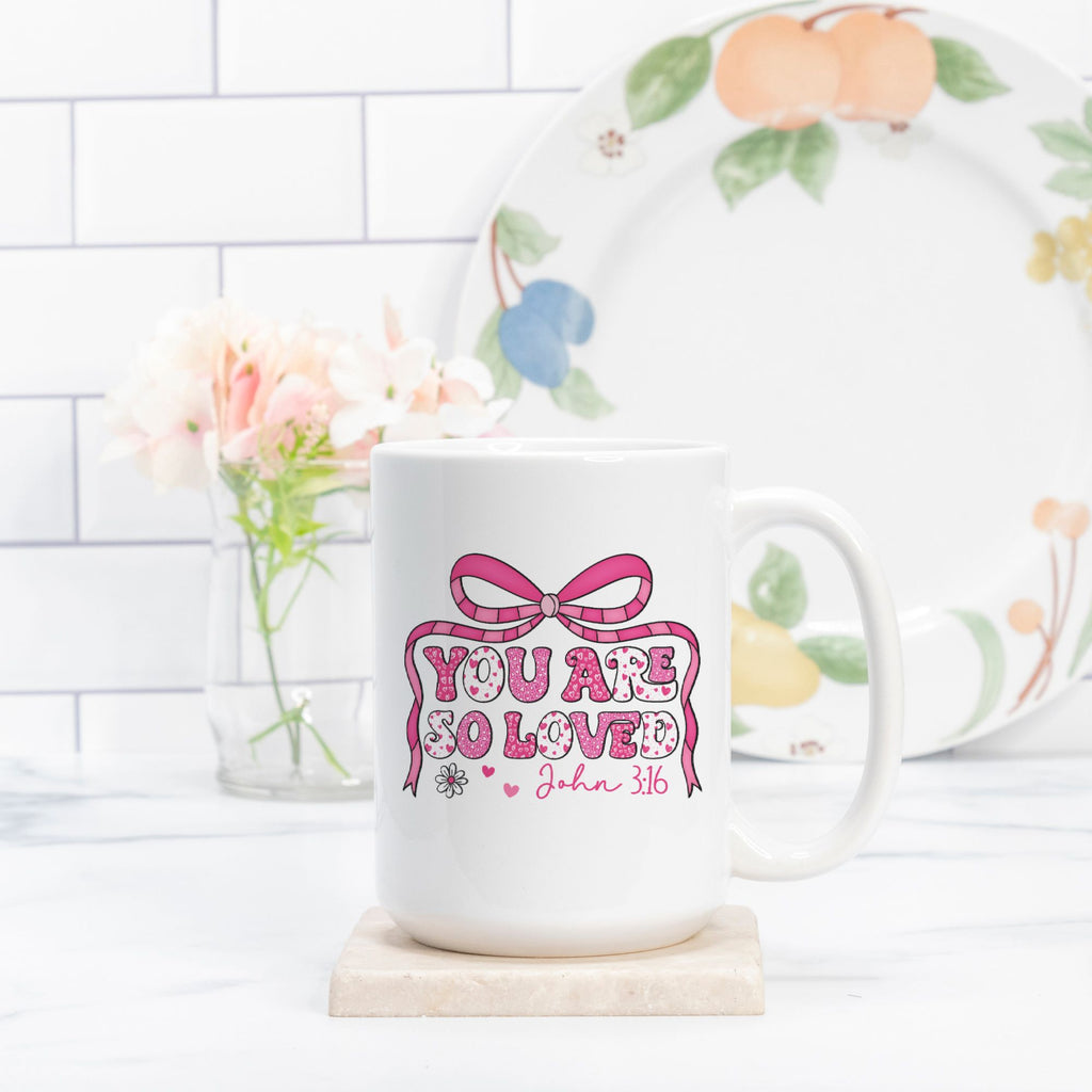White mug with 'You are so loved' text on a white surface with a decorative plate in the background.