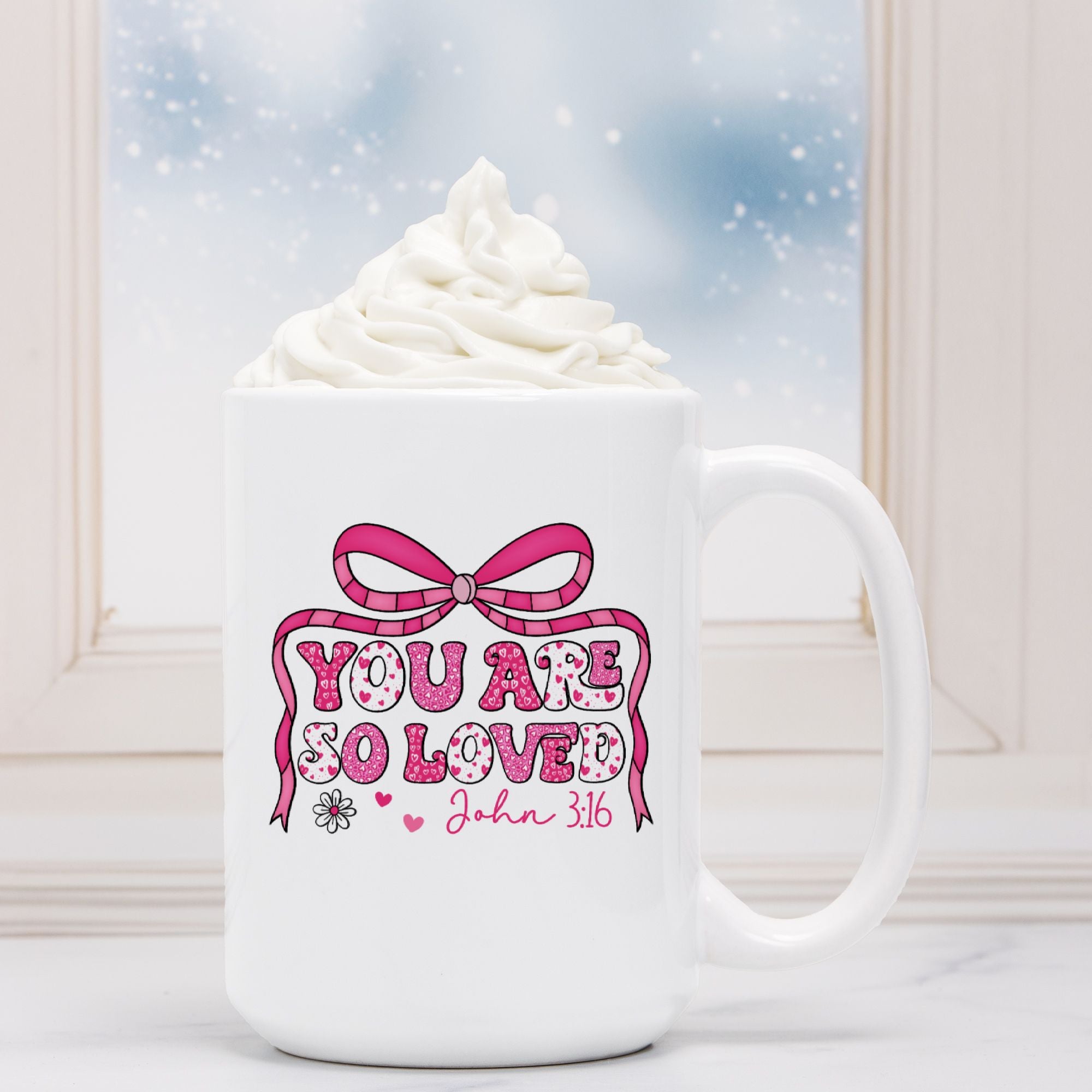 White mug with pink decorative text and bow design, placed in front of a hot chocolate with whipped cream.