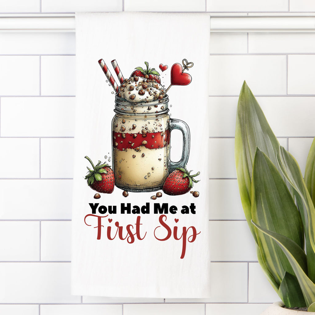 Tea towel with Illustration of a mason jar with a milkshake, strawberries, and heart-shaped straws and text ' You Had Me At First Sip' on a white tiled background.