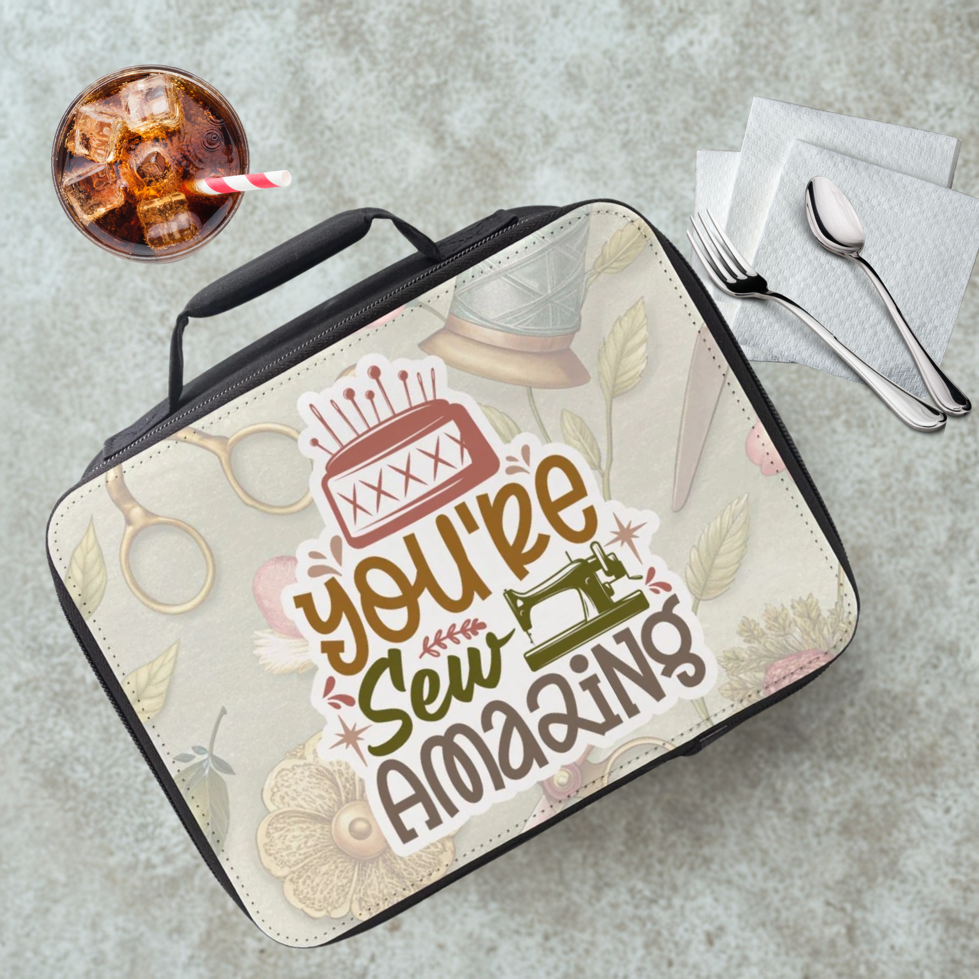 Lunch bag with 'You're Sew Amazing' text on a gray cafeteria table with a drink and utensils.