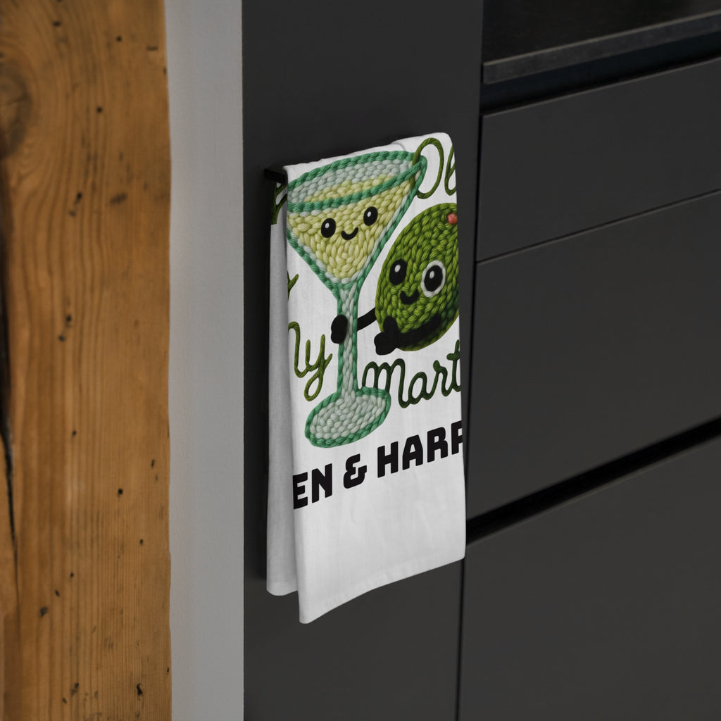 Tea towel with a design of a martini glass and olives hanging on a black cabinet.
