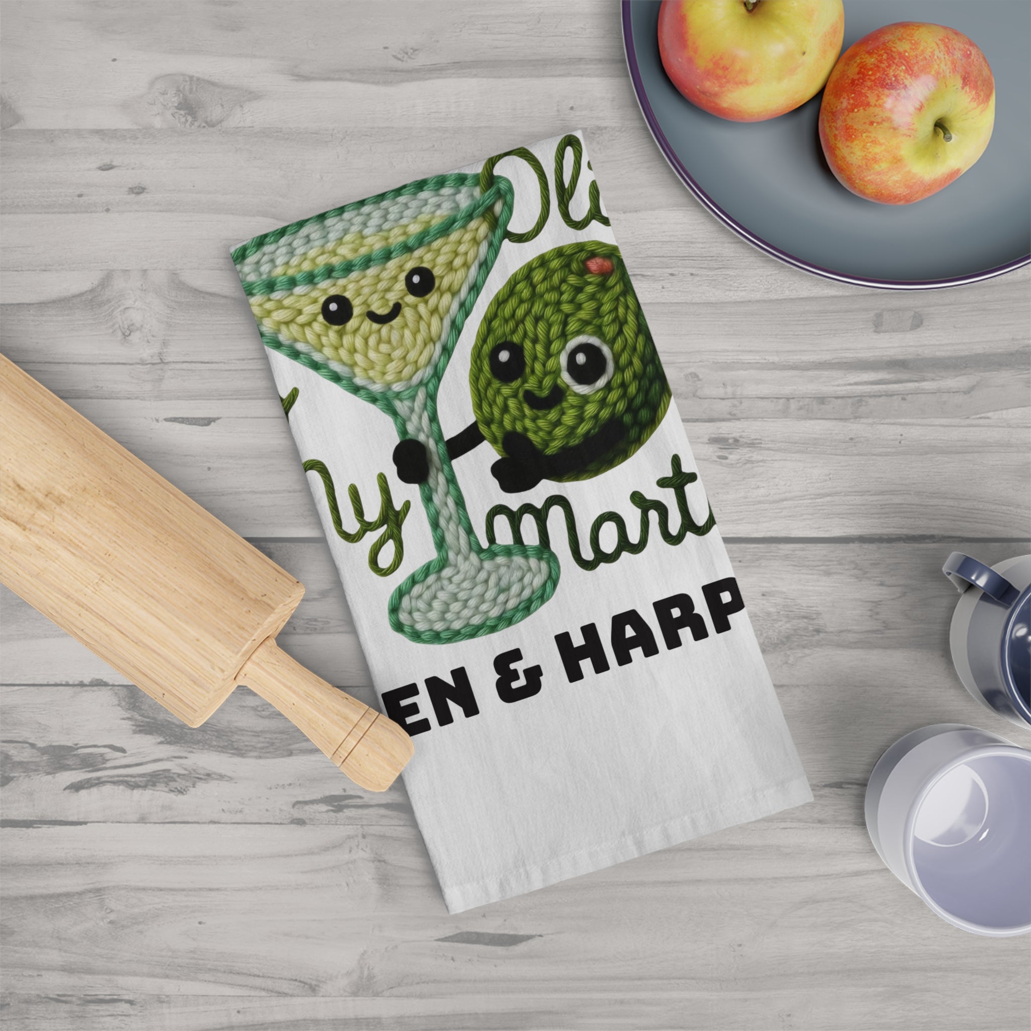 Tea towel with an olive and martini glass design on a kitchen counter with apples and a rolling pin.