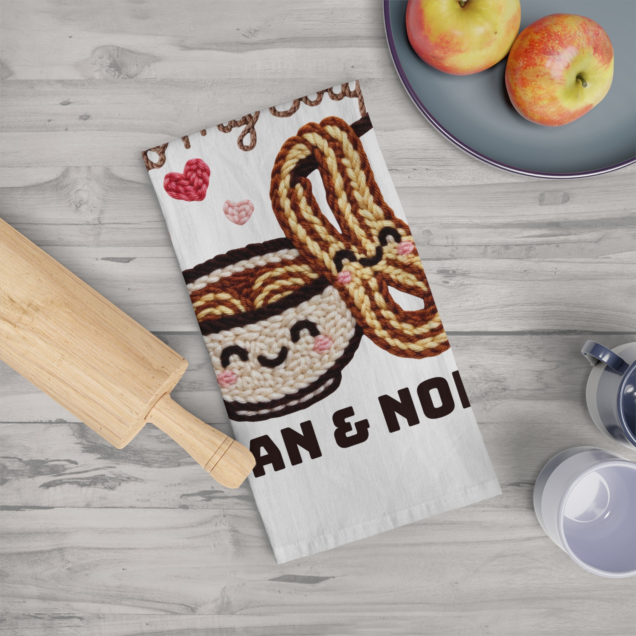 Decorative kitchen towel with a design of noodles and text on a wooden surface with apples and a rolling pin.