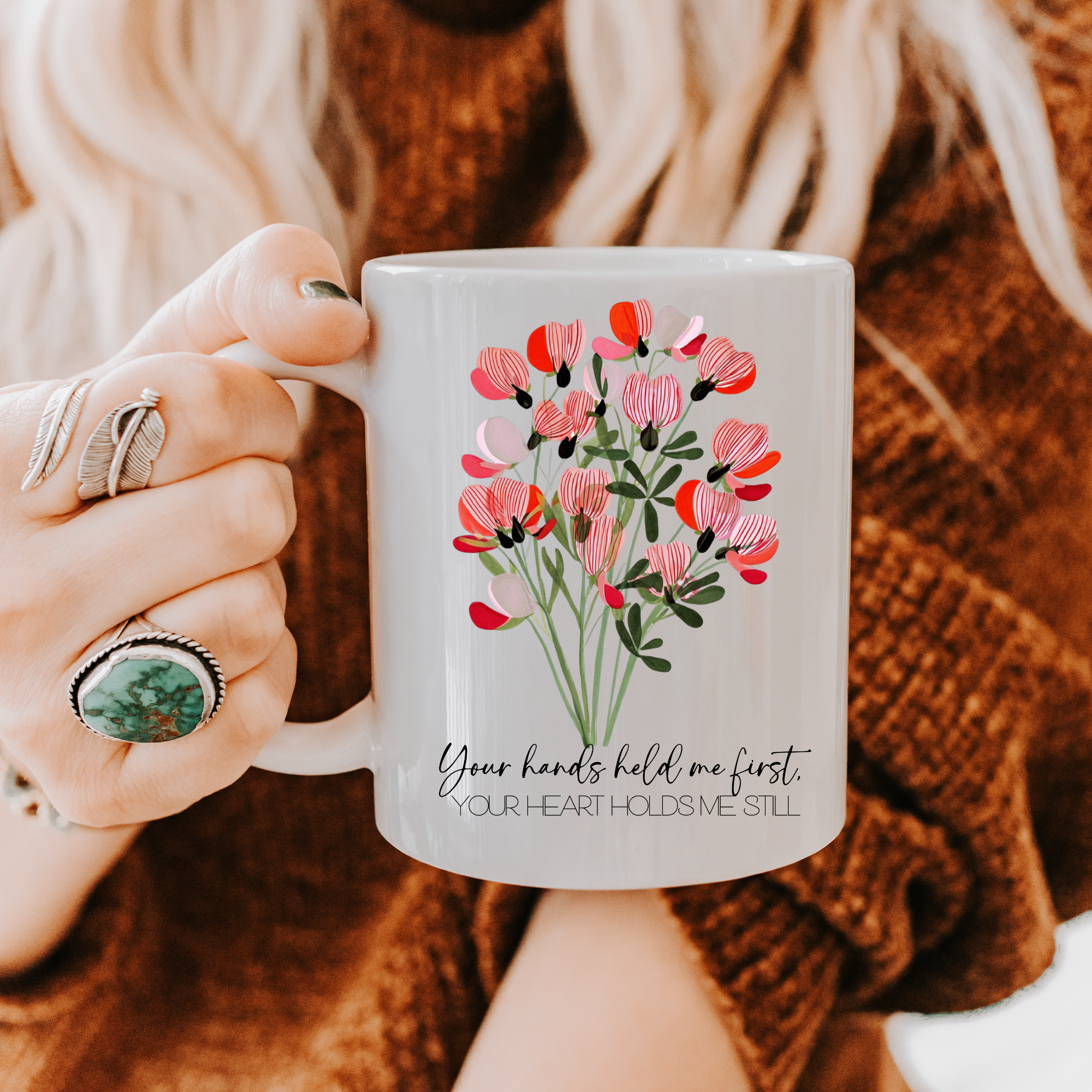 Person holding an 11oz mug with floral design and text, wearing a brown sweater.