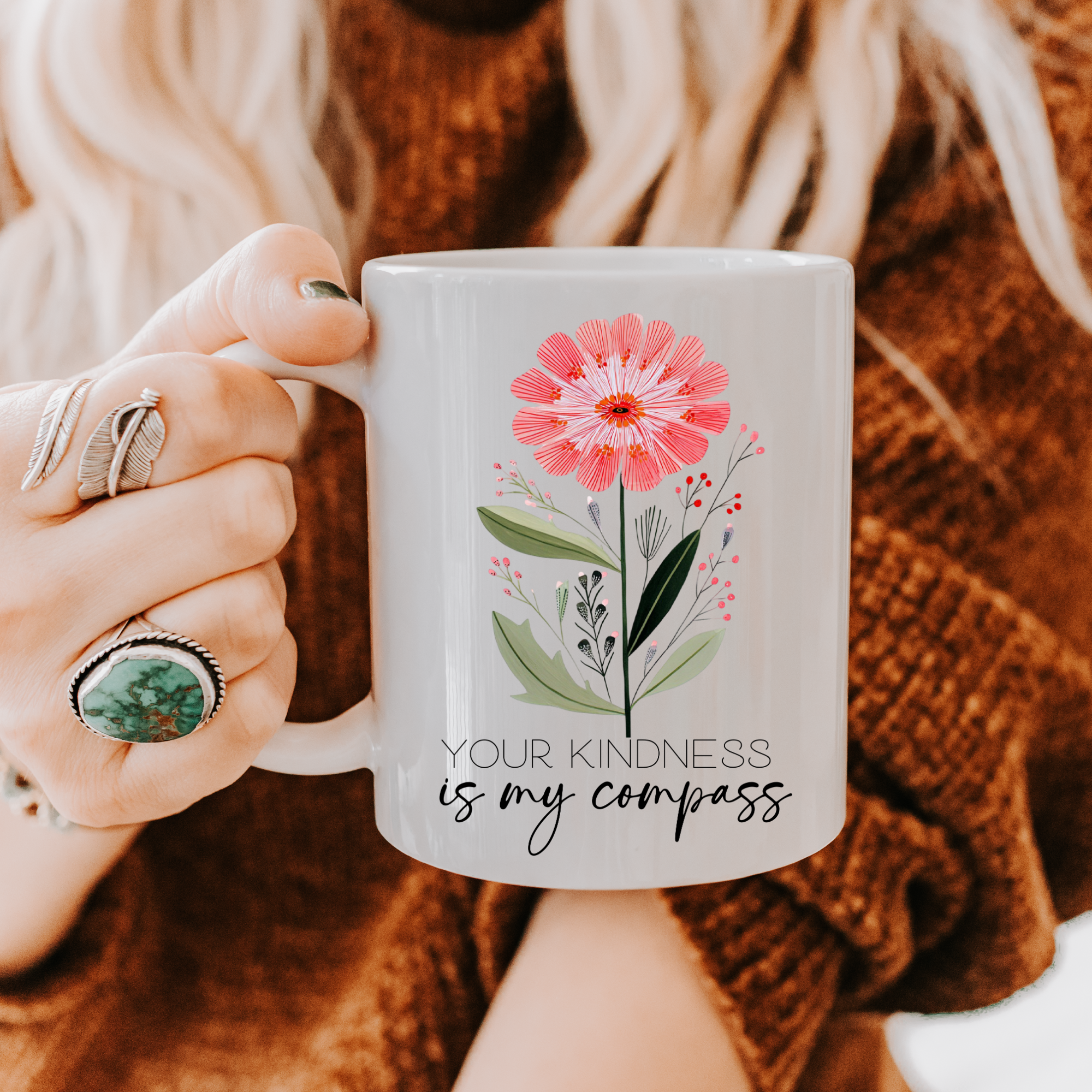 Person holding an 11oz mug with a floral design and text 'Your kindness is my compass' against a blurred background.
