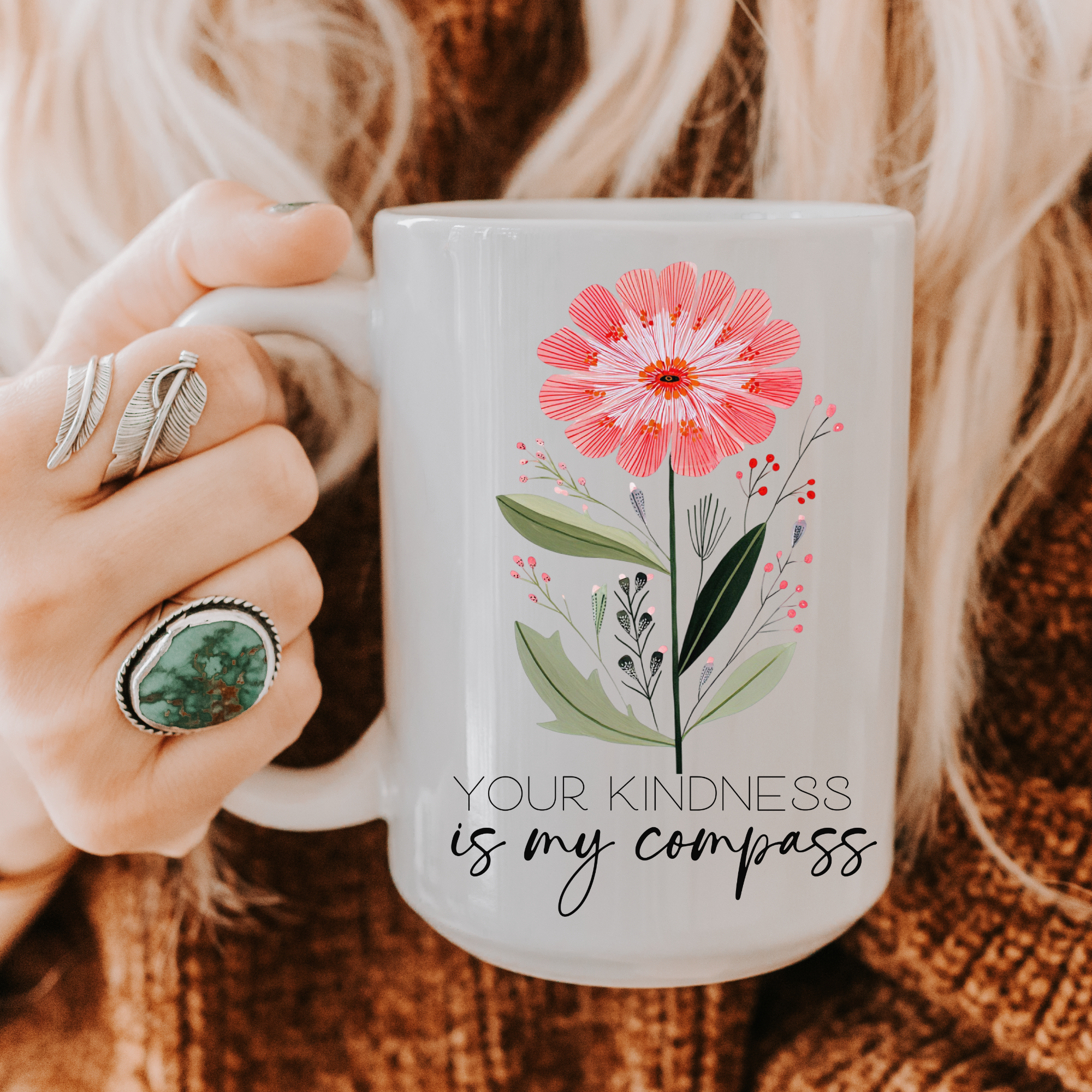 Person holding a 15oz mug with floral design and text 'Your kindness is my compass' on a textured fabric background.