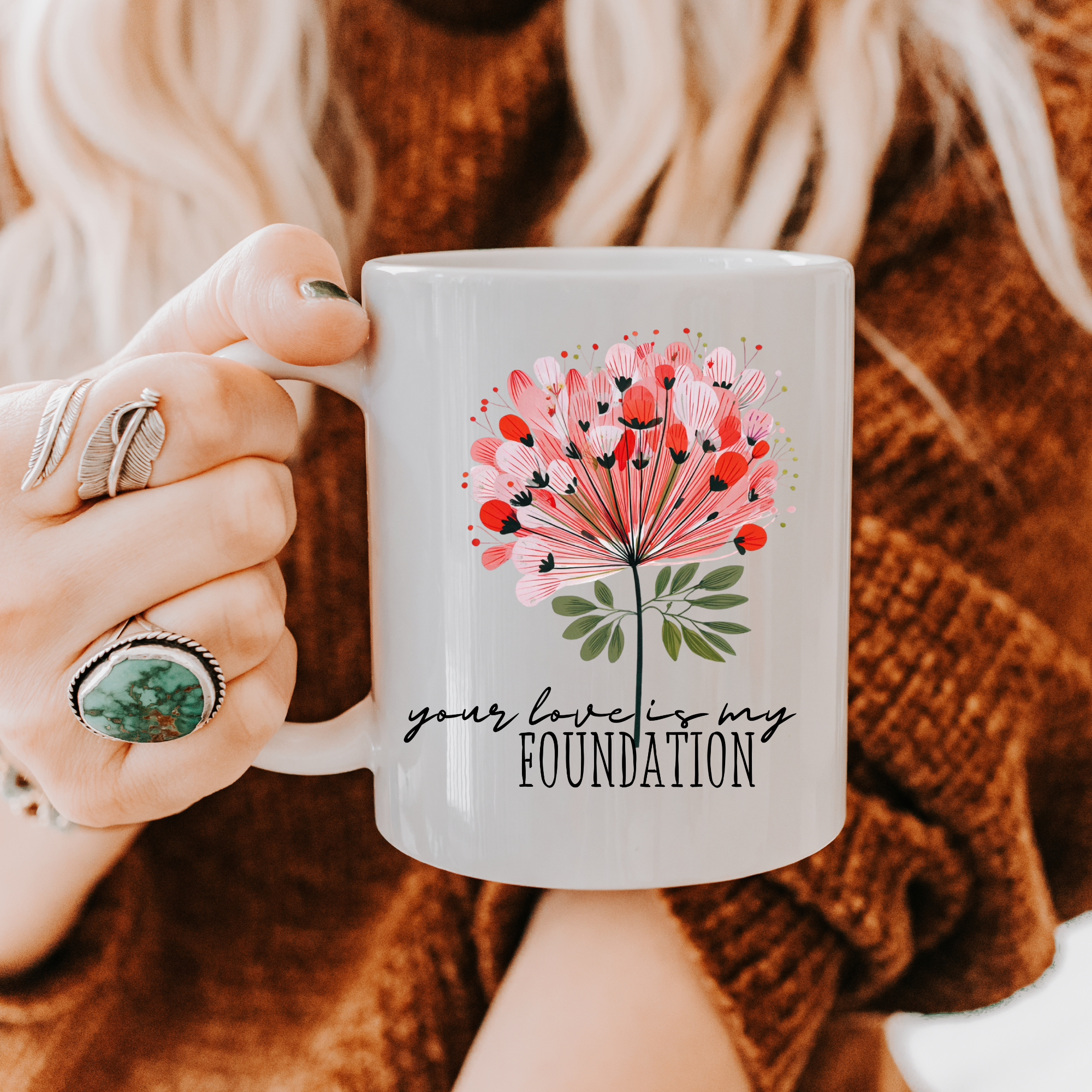 Person holding an 11oz white ceramic mug with a floral design and text 'your love is my foundation' against a blurred background.