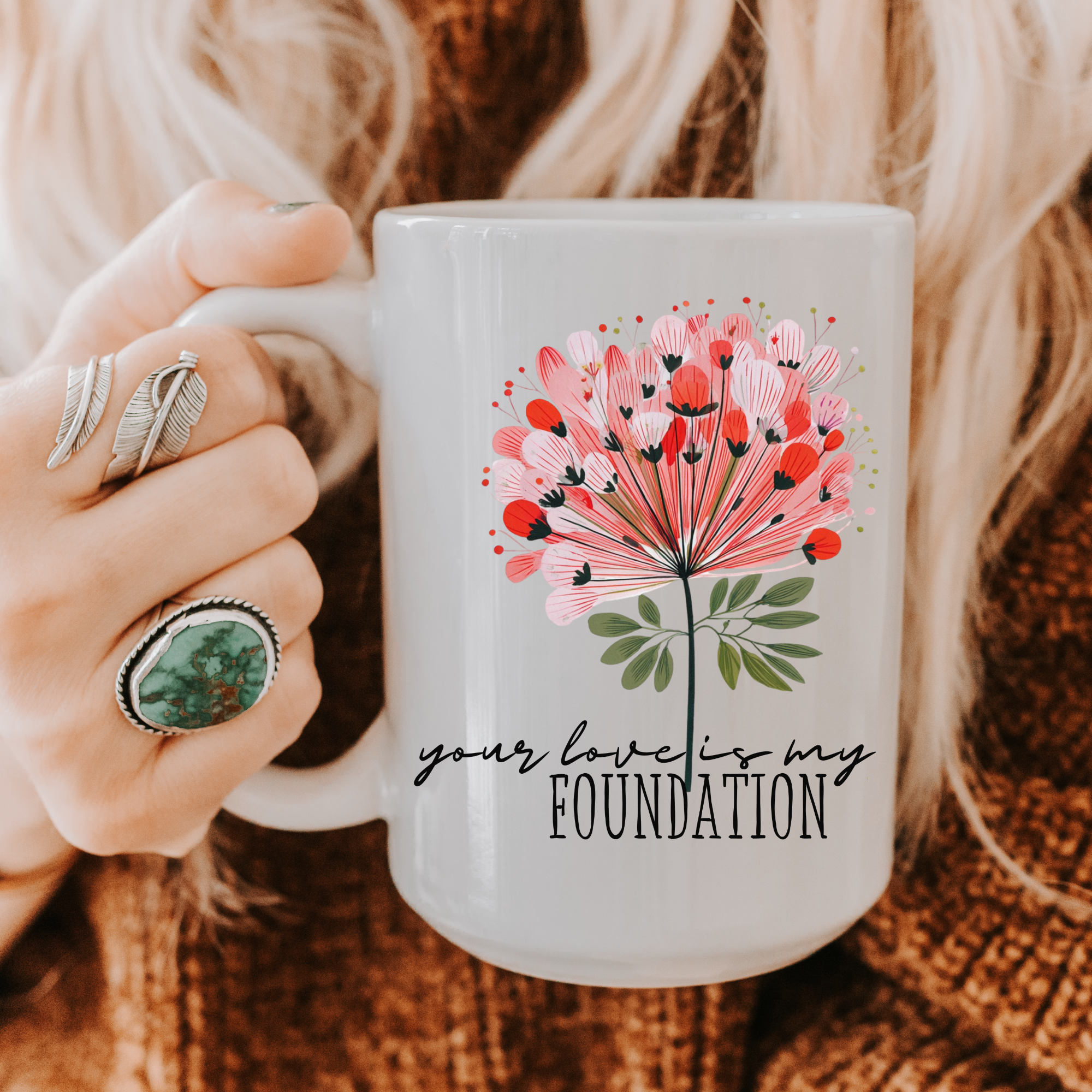 Person holding a 15oz white ceramic mug with a floral design and text 'your love is my foundation' on a textured surface.