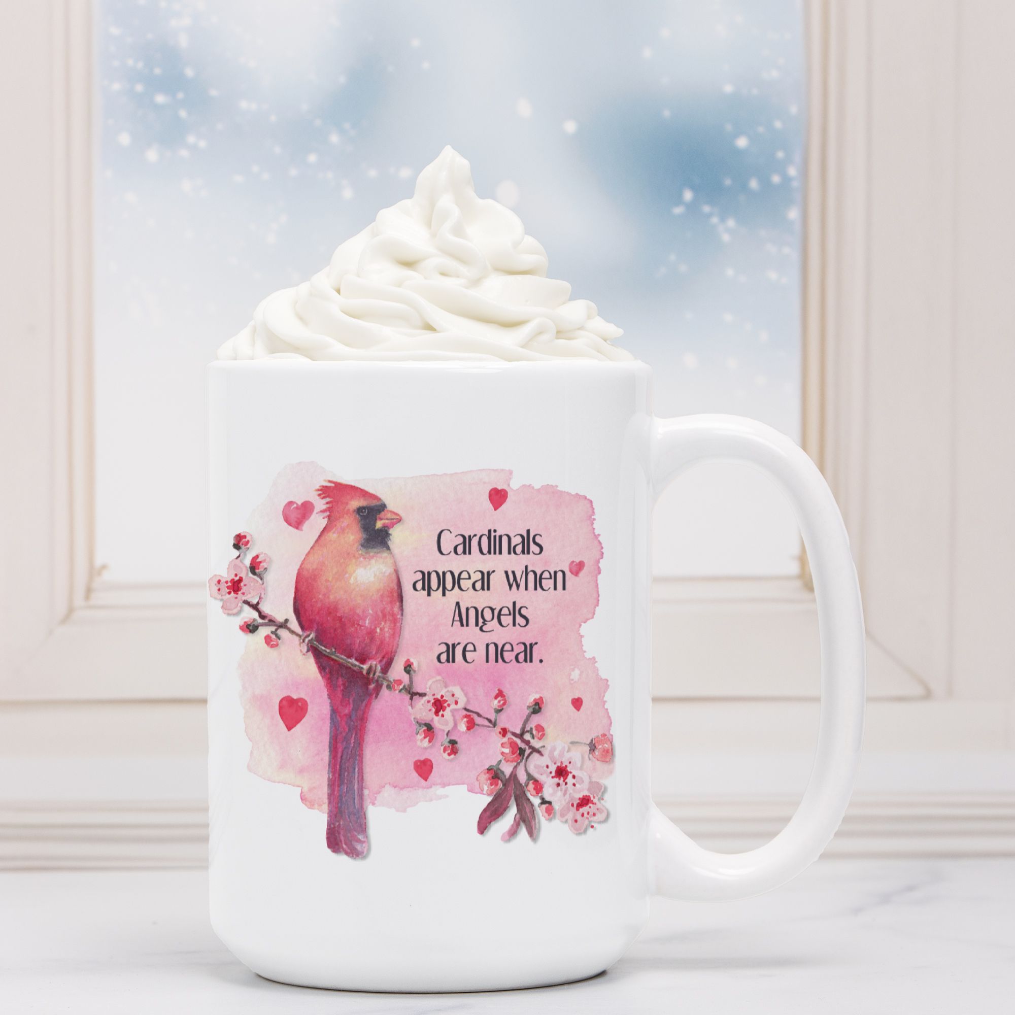 White mug with a pink cardinal design and text, filled with hot chocolate and whipped cream.
